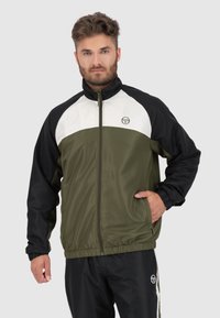 Giacca a vento verde oliva e nera con un pannello superiore bianco, zip frontale, polsini elasticizzati e tasche laterali. Presenta un logo sul petto.