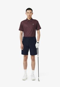 Burgundowy polo w wzory zestawiony z granatowymi szortami. Model trzyma kij golfowy, nosi białą rękawiczkę i sneakersy. Prosty, sportowy design.