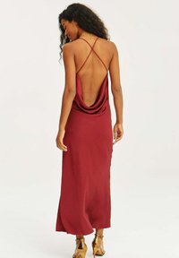 Bordová saténová maxi šaty s obojkom vzadu, tenkými prekríženými ramienkami a bočnými rozparkami. Modelka má na sebe zlaté vysoké topánky.