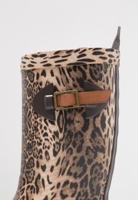 Botte en caoutchouc à motif léopard avec un accent en cuir marron, un large revers et des composants en plastique noir. Surface texturée avec des couleurs contrastées.