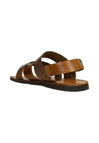Sandale en cuir marron avec une semelle plate, présentant des brides croisées et un accent texturé. Le design est ouvert au bout et a une forme minimaliste.