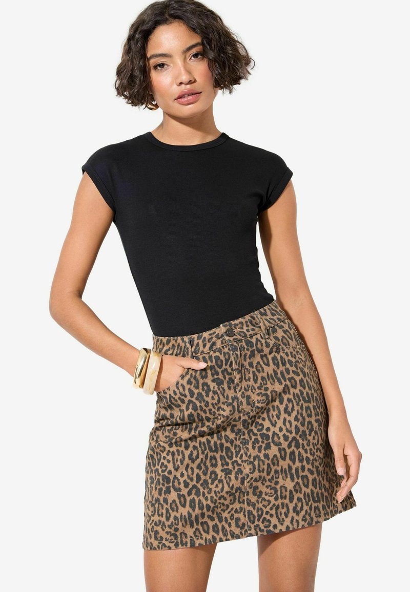 Zwarte korte mouwen top gecombineerd met een bruine denim rok met luipaardprint, voorzien van zakken en een knoopsluiting aan de voorkant. Grote armbanden worden gedragen.