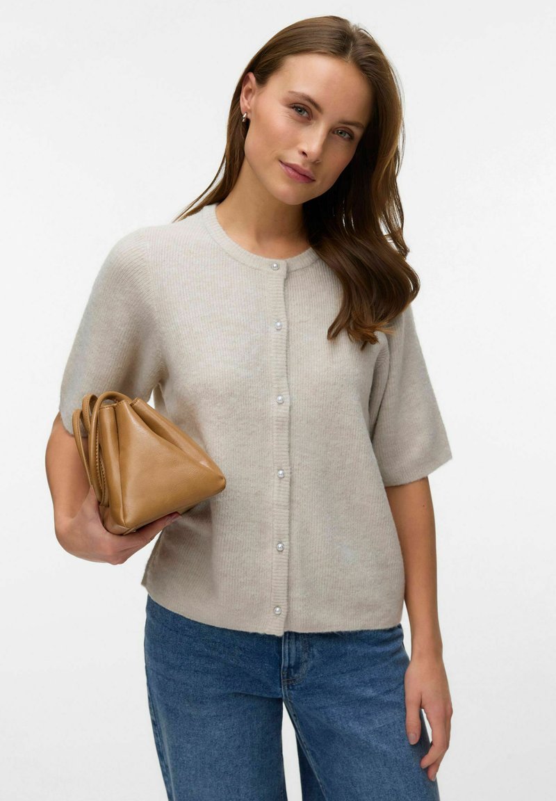 Femme aux longs cheveux bruns portant un pull beige boutonné et un jean bleu, tenant un sac à main en cuir beige sur fond blanc.