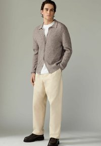 Cardigan gri cu pliuri, butonat în față, purtat peste o cămașă albă. Asortat cu pantaloni lejeri de culoare crem și pantofi negri cu tălpi texturate.