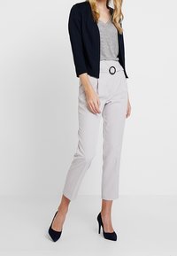 Kvinna som bär en svart kort kavaj, randig V-ringad topp, ljusgrå byxor med bälte och svarta högklackade skor mot vit bakgrund.