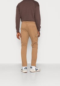 Khaki cargo kalhoty s mnoha kapsami, gumovým pasem a zúženým střihem. Model má na sobě hnědou mikinu a tenisky.