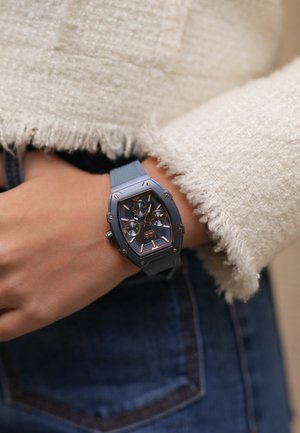 Montre avec un bracelet en caoutchouc bleu, un boîtier hexagonal, des accents dorés et plusieurs sous-cadrans. Portée sur un poignet, avec un pull texturé en arrière-plan.