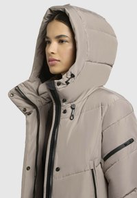 Chaqueta tipo puff en beige claro con capucha, que cuenta con cierre de cremallera, botones de presión, bolsillos laterales y una textura suave y acolchada.
