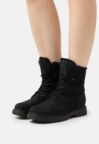 Bottes bottines en suède noir avec doublure en fourrure, devant à lacets et semelle en caoutchouc. Caractéristiques : bout rond et boutons décoratifs sur le côté.