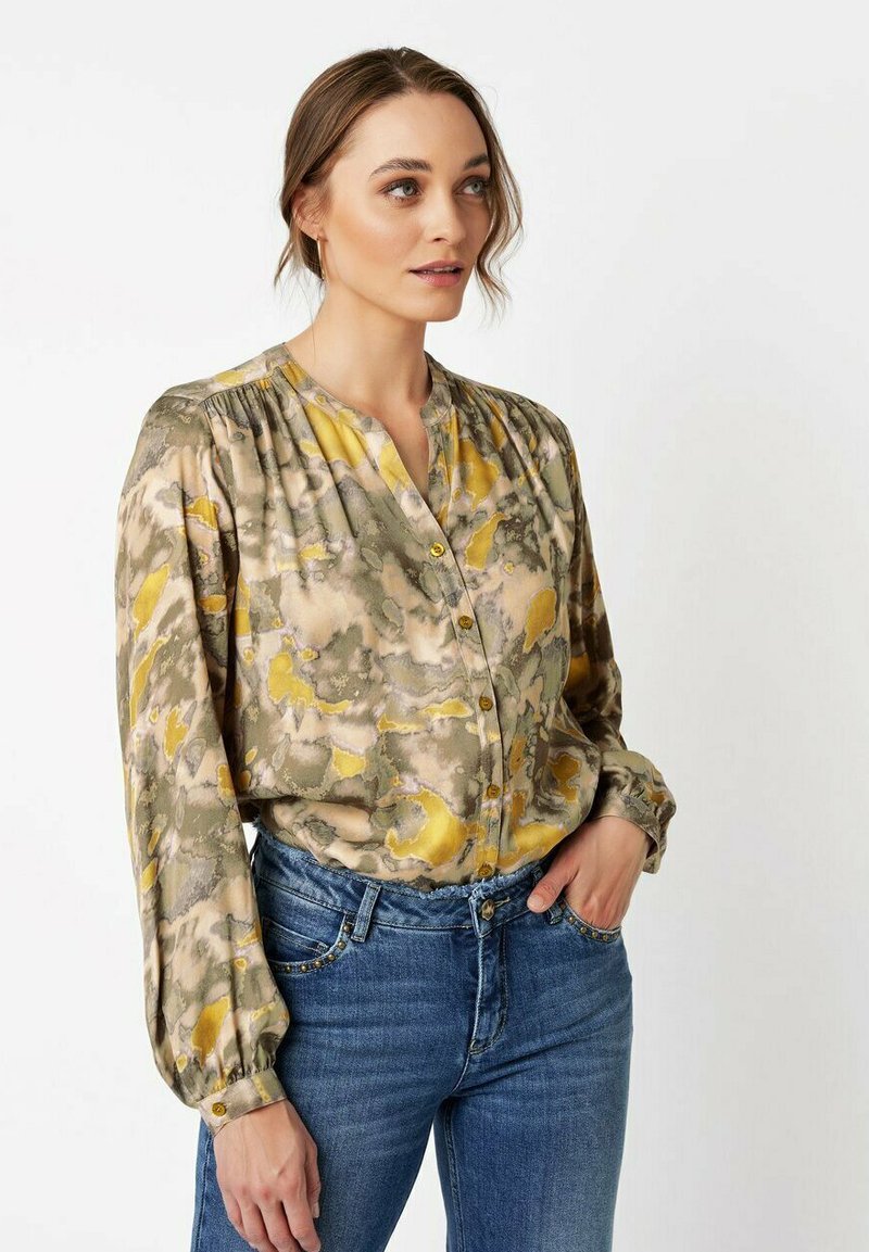 Frau trägt eine lockere, langärmlige Bluse mit Knöpfen im gelb-beigen, abstrakten Muster, kombiniert mit blauen Jeans, stehend vor einem weißen Hintergrund.