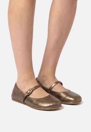 Beine in bronzefarbenen Mary Jane Ballerinas mit einem dünnen Schnallenriemen vor hellgrauem Hintergrund.