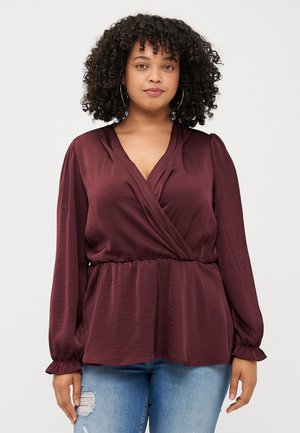A woman with curly hair wearing a burgundy wrap top and blue jeans stands against a light background, looking directly at the viewer.