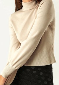 Schlanker beiger Rollkragenpullover mit gerippten Bündchen und Saum, der eine glatte Textur aufweist. Kombiniert mit einem strukturierten schwarzen Rock.