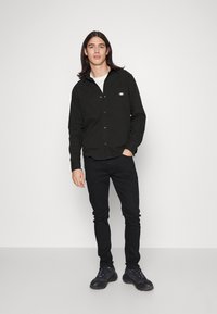 Camisa negra de botones hecha de tela, con un parche de logo en el bolsillo del pecho, combinada con jeans negros ajustados y zapatillas oscuras.