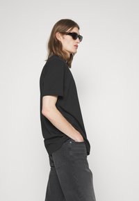 T-shirt en coton noir à manches courtes, associé à un jean gris foncé. Le modèle porte des lunettes de soleil noires, posé de profil contre un fond clair.