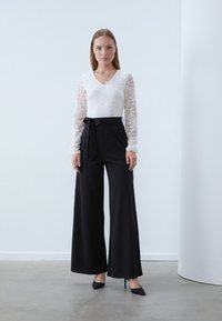 White lace long-sleeve blouse with a V-neck, paired with high-waisted black wide-leg trousers featuring a bow tie at the waist. Heeled shoes.
