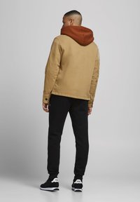 Giacca di cotone beige con cappuccio marrone, pantaloni felpa neri e sneaker nere. La giacca presenta aperture laterali e polsini con bottoni.
