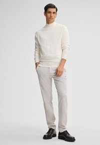 Maglione bianco a maglia con collo alto, abbinato a pantaloni beige chiaro e scarpe nere con lacci, caratterizzato da texture lisce e design aderente.