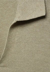 Beige geribbelde gebreide stof met een licht reliëf, voorzien van een geplooide rand. De stof is zacht en rekbaar, geschikt voor diverse ontwerpen.