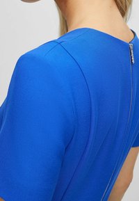 Niebieska sukienka z krótkim rękawem, uszyta z gładkiego, teksturowanego materiału. Posiada delikatny detal szwu oraz zamek błyskawiczny z tyłu.