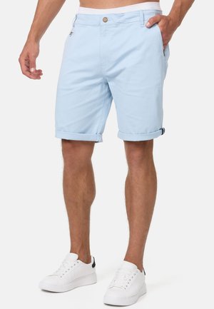 Hellblaue Baumwollshorts mit umgeschlagenen Bündchen, Seitentaschen und einem vorderen Knopfverschluss. Kombiniert mit weißen Sneakers mit schwarzem Akzent.