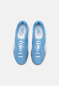 Sapatilhas azul-sedad com detalhes brancos e atacadores azul-claro. Apresentam um bico arredondado e uma sola em borracha castanha texturizada. Logotipo da Reebok na língua.