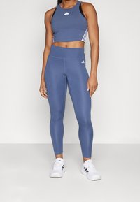Blå atletisk crop top med tre vita ränder på sidorna, kombinerad med blå tights med hög midja och logotypdetaljer. Vita sneakers.