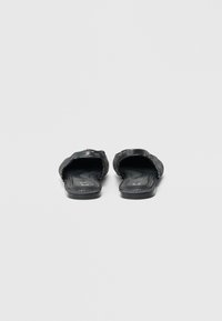 Černé nazouvací sandály s hladkým povrchem, zdobené kovovými cvočky podél popruhů a okrajů, otevřenou špičkou a plochou podrážkou.