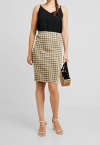 Černý bezramínkový top, žlutá kostkovaná tužková sukně a béžové sandály na kotníku. Modelka drží malou, tkanou černou kabelku.