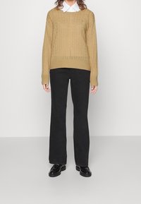 Khaki-Strickpullover mit breitem Ausschnitt, kombiniert mit schwarzen ausgestellten Hosen und schwarzen glänzenden Loafers. Darunter ist ein weißes Hemd mit Kragen sichtbar.