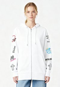 Bela jopa z zadrgo, na rokavih z grafičnimi in besedilnimi motivi, vključno z barvitimi dizajni in tiskanimi frazami. Izdelana iz mehkega blaga.