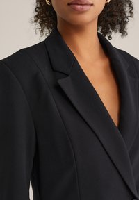 Blazer negru cu decolteu în V adânc, rever structurat și textură netedă. Prezintă umeri adaptați și siluetă ajustată.