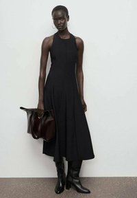 Vestido negro sin mangas con falda acampanada, combinado con botas negras hasta la rodilla. Sosteniendo un gran bolso marrón oscuro. Textura suave, diseño minimalista.