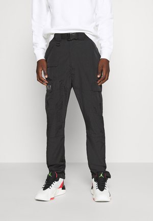 Personne portant un pantalon cargo noir avec plusieurs poches et une ceinture, associé à des baskets montantes blanches, noires, rouges et vertes.