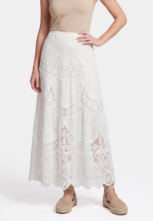 Vrouw met een witte, enkellange kanten rok, beige mouwloos topje en beige espadrilles met enkelbandjes, staand met één hand op de heup.