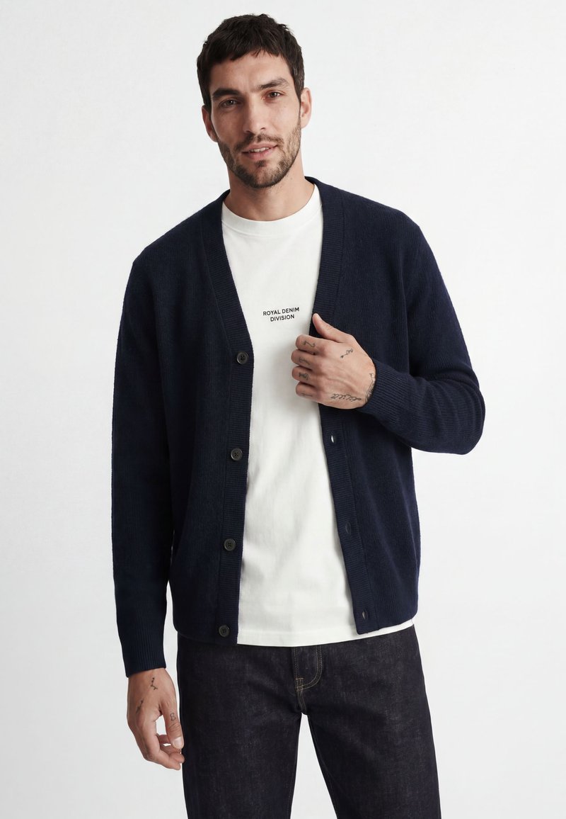 Homme portant un cardigan bleu marine boutonné sur un t-shirt blanc avec le texte "Royal Denim Division" et un jean foncé sur un fond uni.