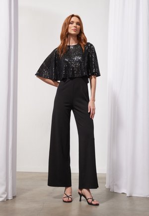 Salopetă neagră cu picioare largi și un corset ajustat, acoperită cu o capa scurtă cu paiete. Include pantofi cu toc subțiri, negri, și o textură de țesătură netedă.