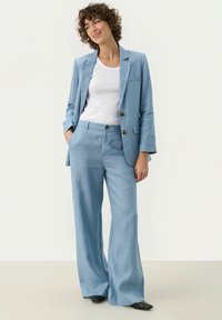 Lichtblauw linnen pak met een getailleerde blazer en wijde broek. De blazer heeft twee knopen en een enkele borstzak. Gemaakt over een witte tanktop.