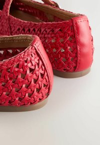 Piros fonott bőrcipők zárt sarokkal és barna gumitalppal, hátulnézetben világos felületen.