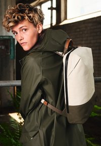 Zaino realizzato in tessuto crema e verde oliva con una forma strutturata. Presenta una cerniera nera, dettagli in pelle e spallacci regolabili.