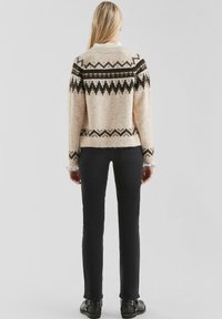 Wollmischpullover in Hellbeige mit schwarzen geometrischen Mustern, Rundhalsausschnitt und geripptem Saum, kombiniert mit schwarzen, figurbetonten Hosen und Ankle Boots.