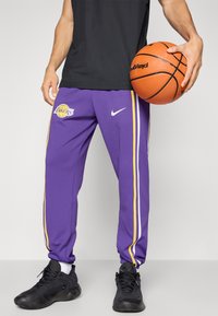 Lilla træningsbukser med hvide og gule sidelister, prydet med et Lakers-logo på venstre lår, parret med sorte sneakers.
