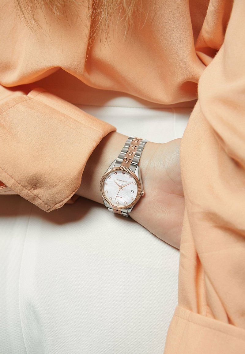 Reloj de pulsera en plata y oro rosa con una esfera redonda de nácar y una pantalla de fecha, que presenta una correa texturizada y marcadores de hora prominentes.