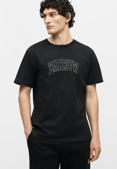 Νεαρός άνδρας με σγουρά μαλλιά που φοράει μαύρο T-shirt με το κείμενο "BARROW" με έντονο λευκό περίγραμμα, τα χέρια στις τσέπες, κοιτάζει προς τα αριστερά.