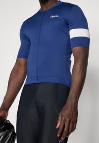Maillot de cyclisme bleu à manches courtes, avec fermeture éclair à l'avant et détails à rayures blanches. Porté avec un short rembourré noir. Tissu lisse et extensible.