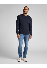 Marinblå sweatshirt med rundad halsringning och ribbade ärmslut, i kombination med ljusblå jeans och vita sneakers. Enkelt varumärke på bröstet.