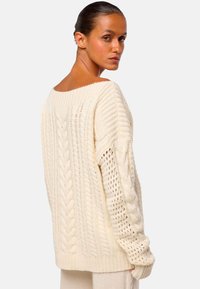 Frau mit nach hinten gekämmtem Haar, die einen cremefarbenen Zopfmuster-Pullover trägt, leicht gedreht und über ihre Schulter blickend, vor einem neutralen Hintergrund.