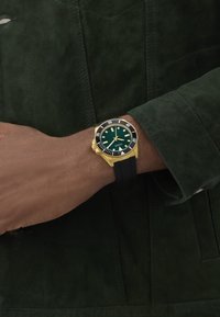 Ceas de scufundare verde cu accente aurii, brățară din cauciuc negru și ace luminescenți. Purtați pe un încheietură, prezentat pe un jachetă din suede verde închis.