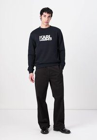 Crna dukserica s bijelim logotipom "KARL LAGERFELD", okrugli izrez, rebrasti rukavi i donji rub. Uparena s širokim crnim hlačama i sjajnim cipelama.