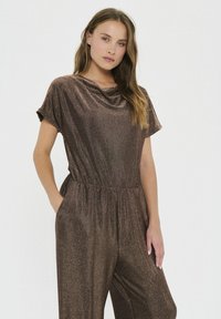 Bronz színű, fémes anyagú kezeslád, rövid ujjakkal, lógó nyakkivágással, gumis derékkal és oldalsó zsebekkel; sima textúrával, amelyen finom csillogás látható.